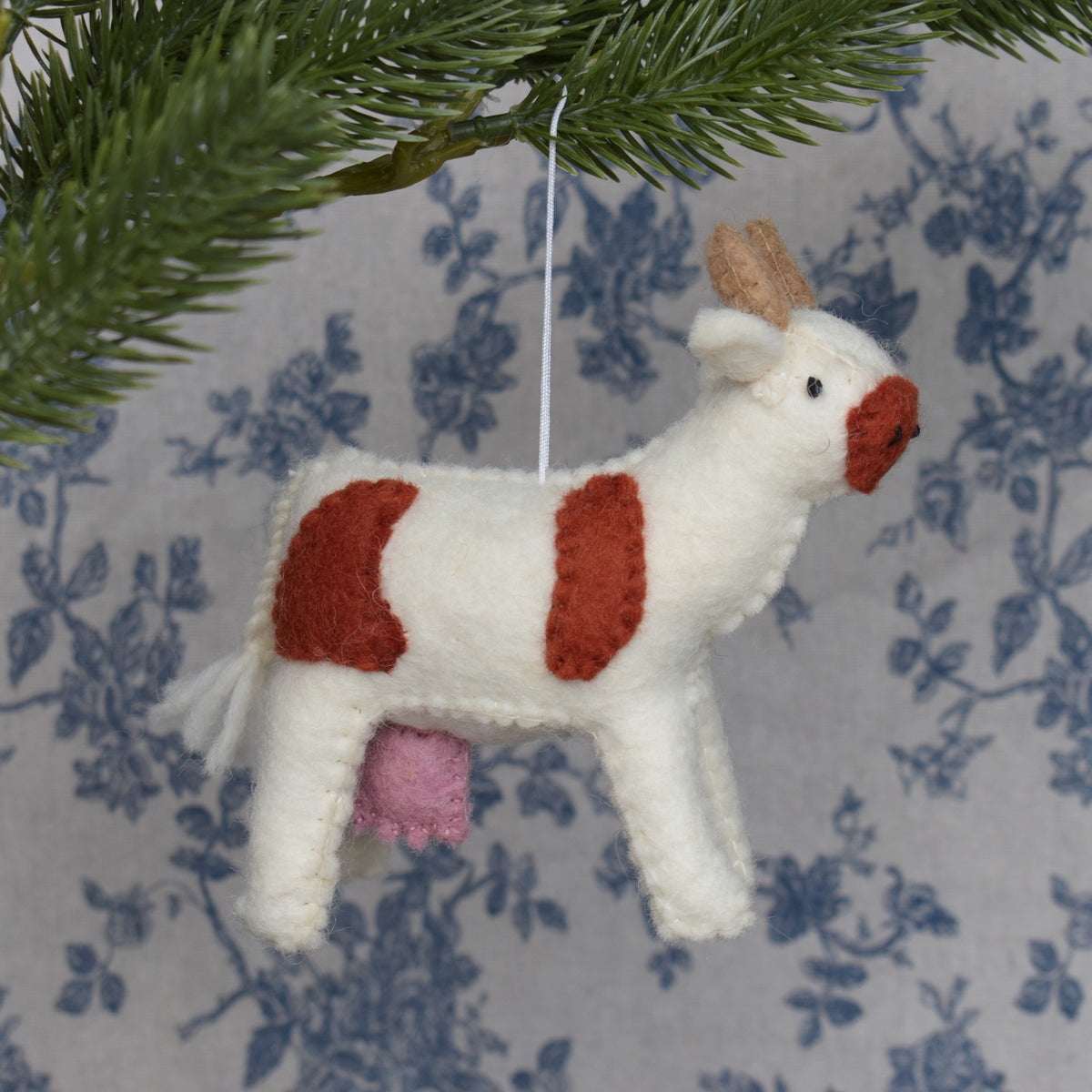 A handmade felt cow Christmas ornament hanging from a pine branch. The ornament is primarily off-white with rust-red patches on its back and head, a small pink felt udder, and light brown felt horns.