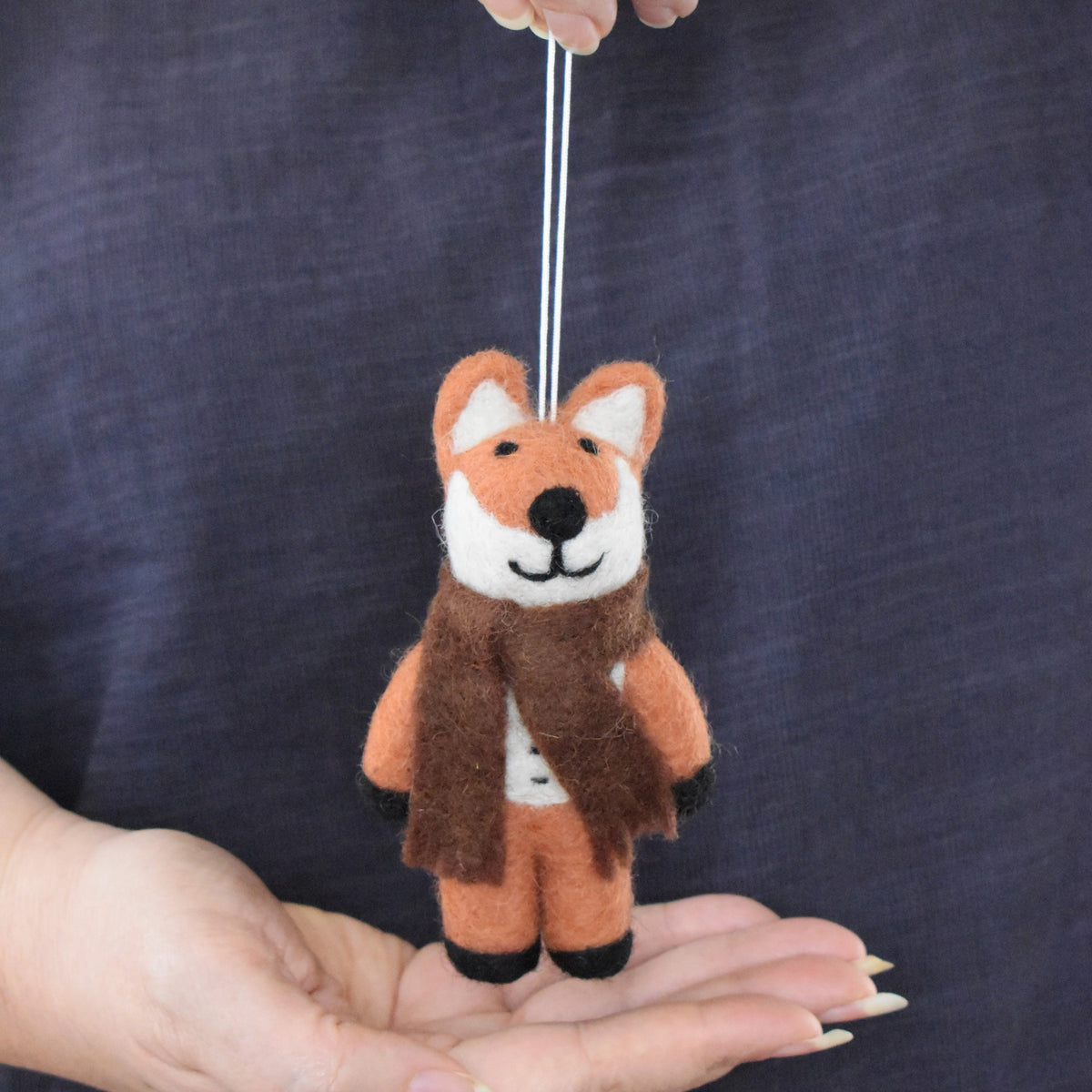 A small felted wool fox ornament wearing a brown scarf, held in a person's hands against a dark navy fabric background.