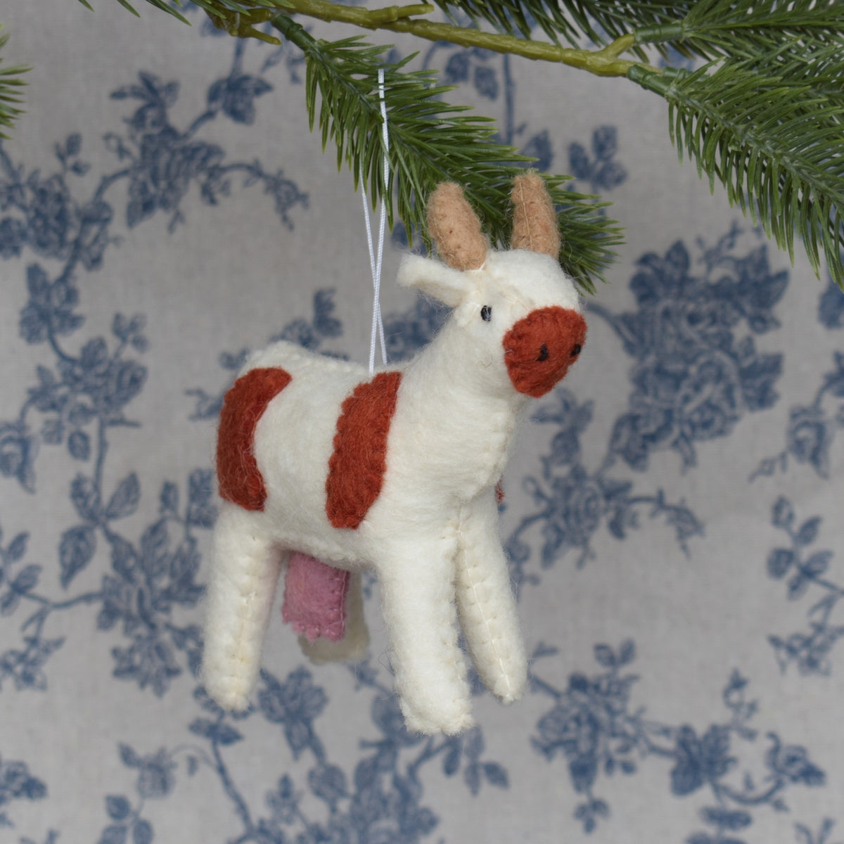 A felt wool cow ornament hanging from a green pine branch against a blue floral patterned background.