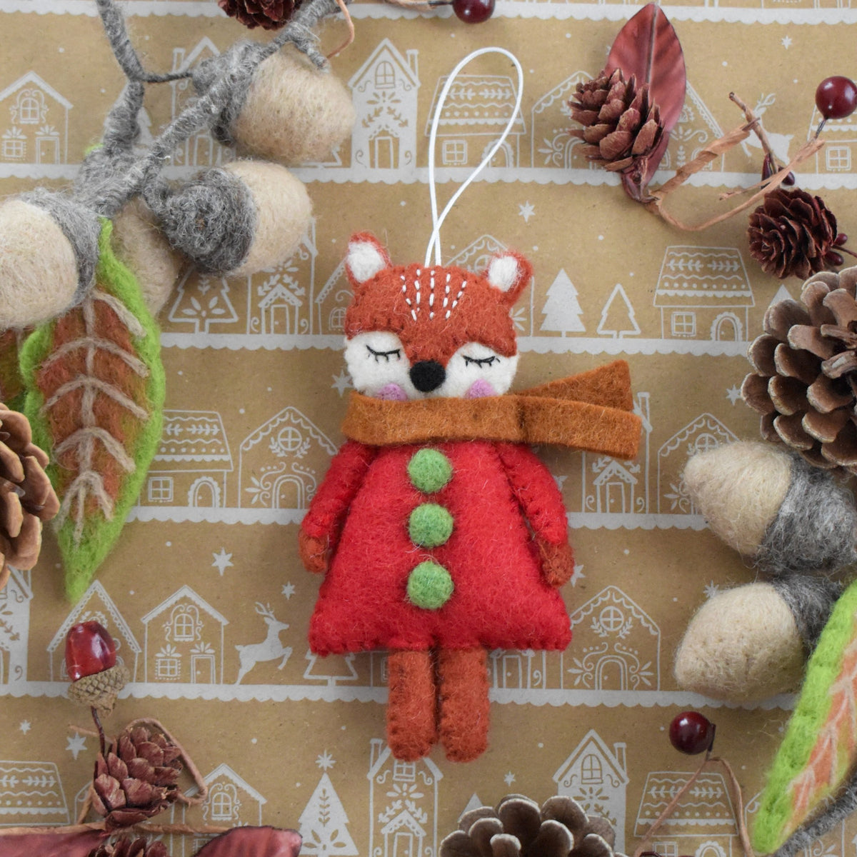 A handmade felt Christmas ornament shaped like a sleeping fox wearing a red coat with green buttons and a brown scarf, hanging against a patterned brown background with pinecones, acorns, and autumn leaves.