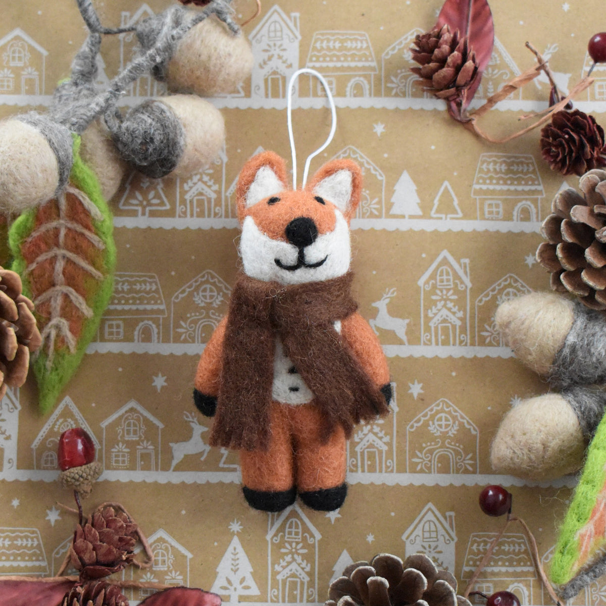 A handmade felt Christmas ornament shaped like a fox wearing a brown scarf, hanging against a decorated kraft paper background with pinecones, acorns, and winter motifs.