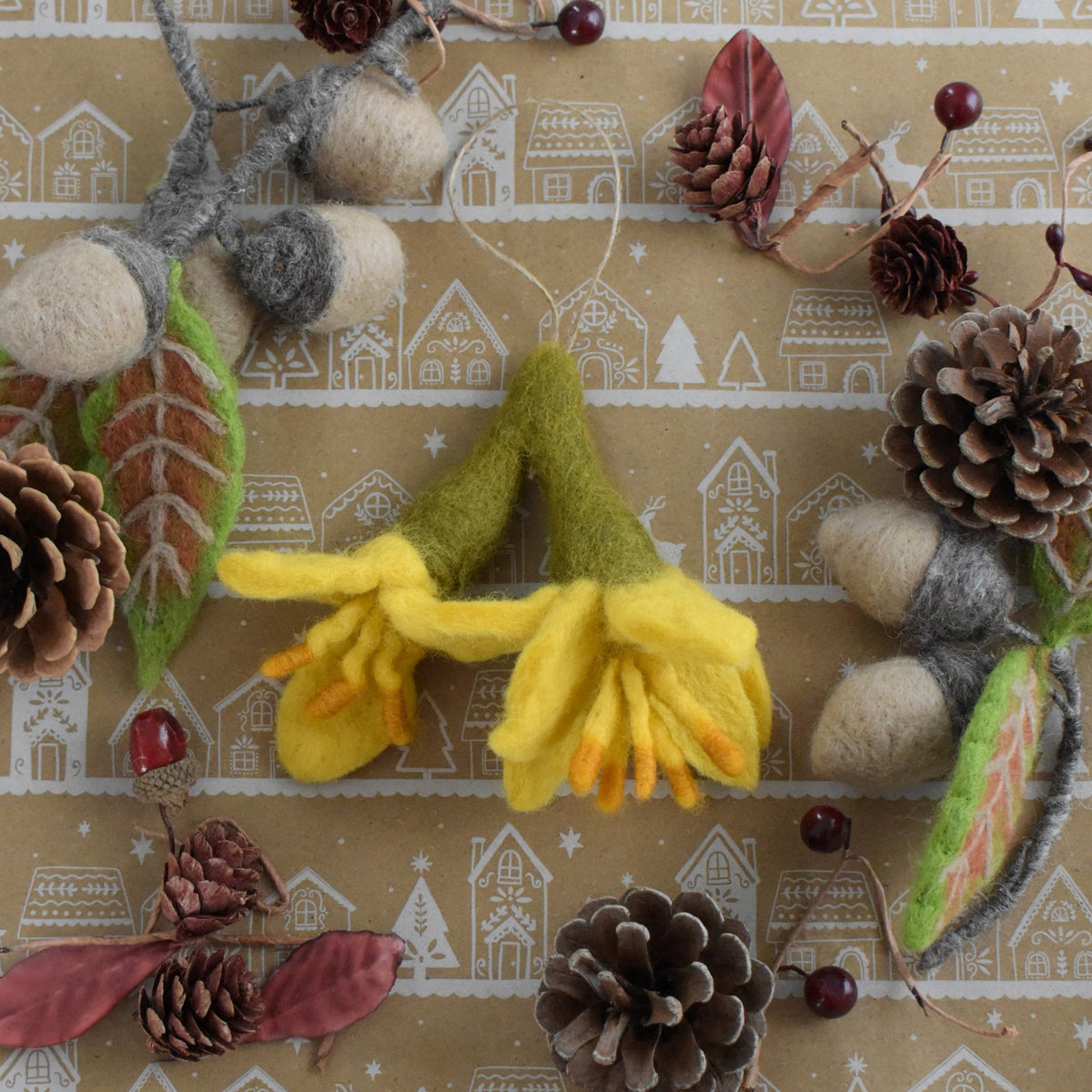 A felted wool hanging ornament shaped like two yellow kowhai with green stems, arranged on a brown kraft paper background printed with a winter village pattern and surrounded by pinecones, dried leaves, and berries.