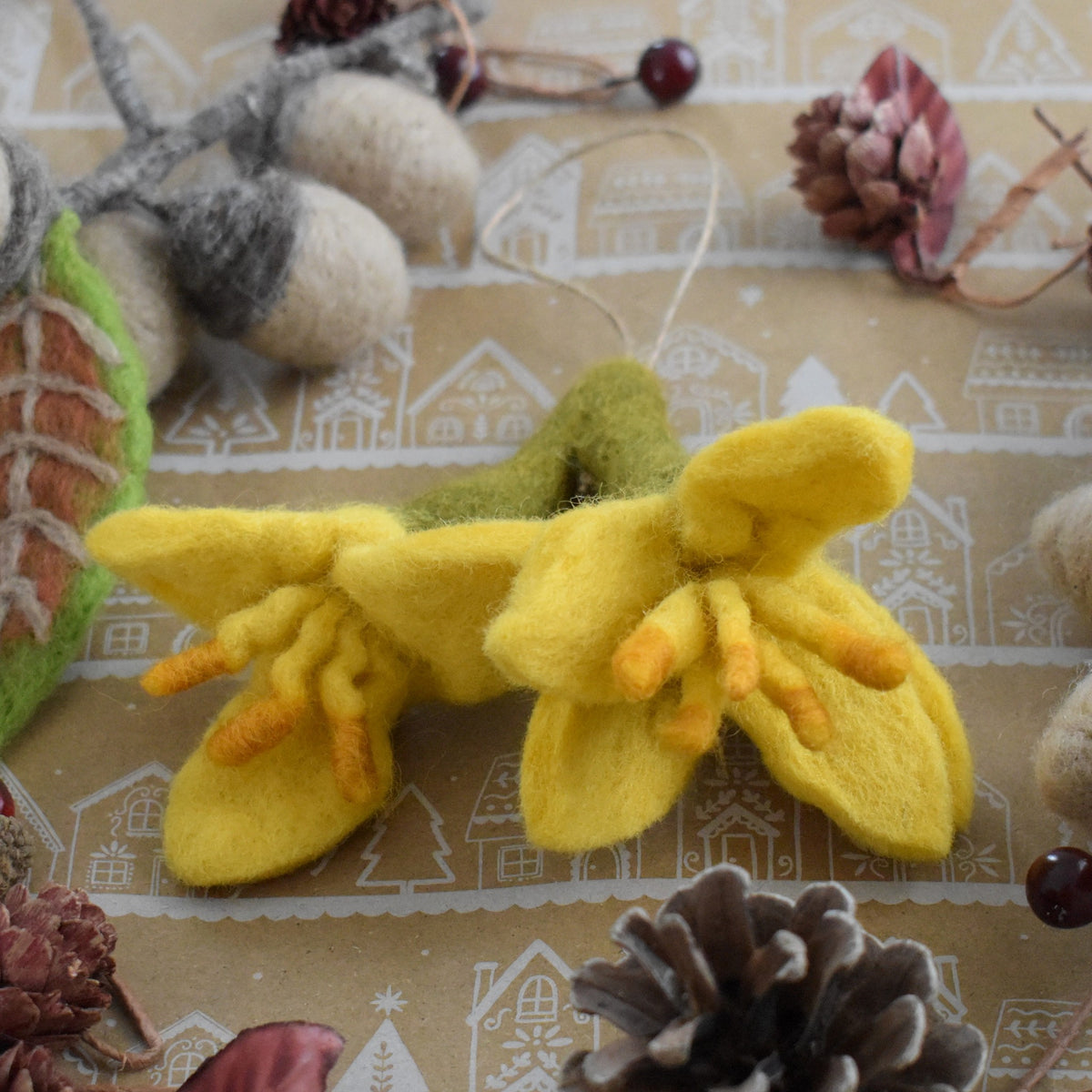 A felted wool ornament depicting two yellow kowhai flowers with orange centers and green stems, resting on a decorated brown background with pinecones and acorns.