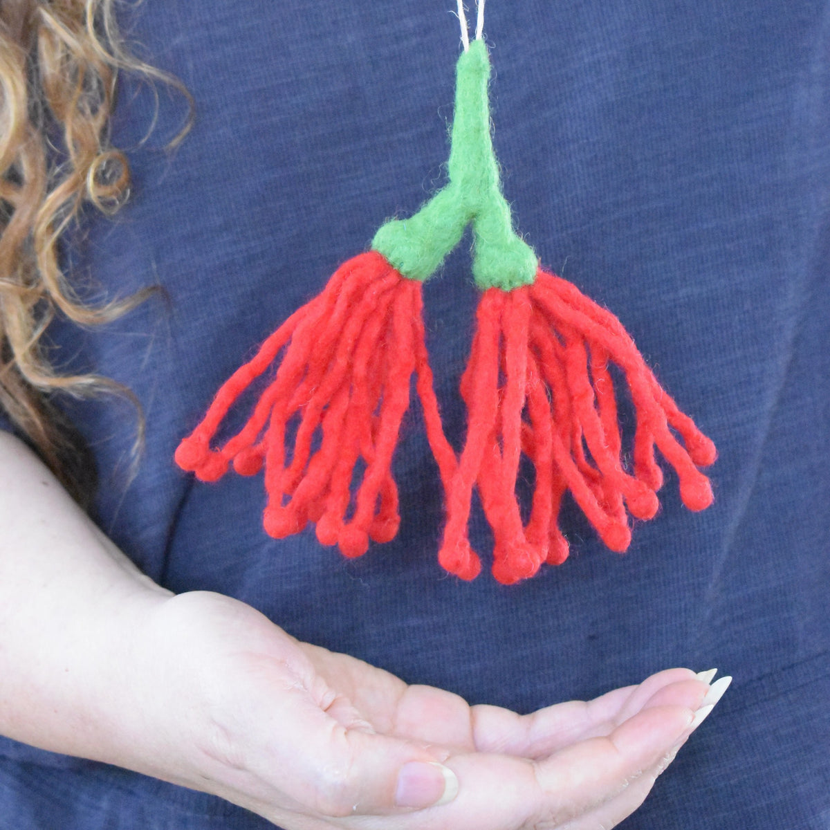 A close-up of a person holding a small felted wool ornament shaped like two red flower-like fronds with green stems, against a dark blue shirt background.
