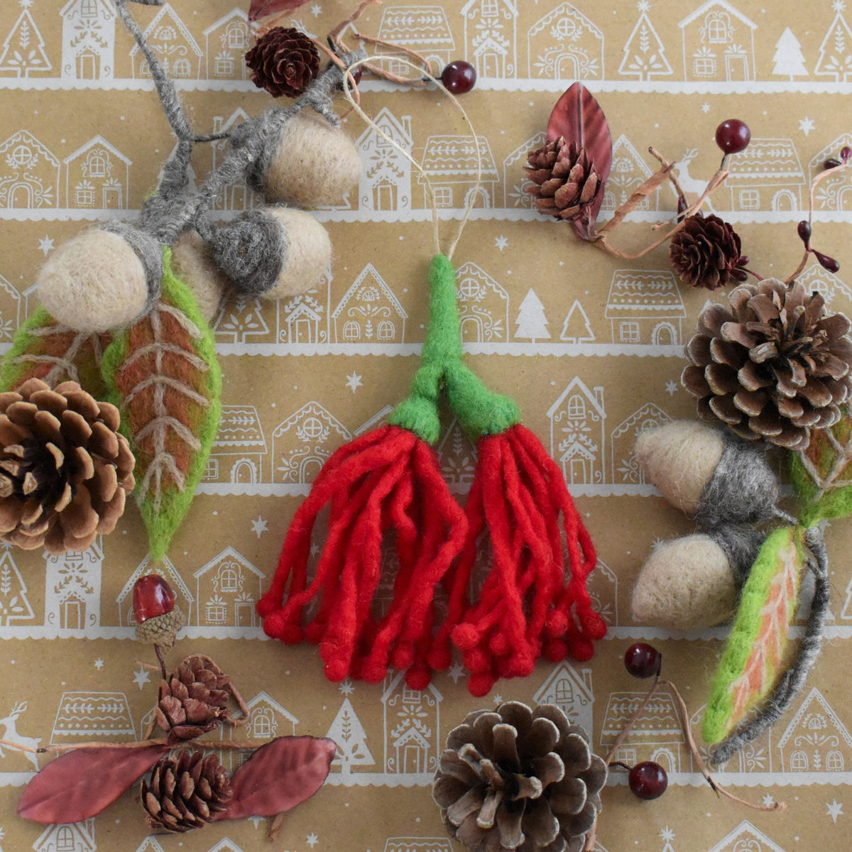 A festive felted wool holiday ornament set featuring red berry sprigs with green tops, pinecones, acorns, and a small bird, arranged on a beige patterned background.